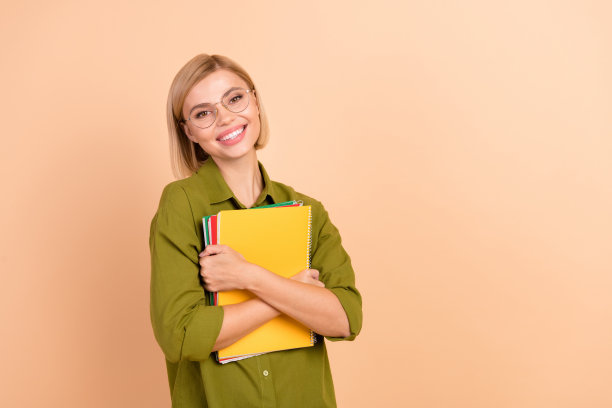 照片中，一个穿着绿色衬衫、戴着眼镜、长着牙齿、笑容灿烂的聪明女人，在米色的背景上拿着备考的课本图片下载