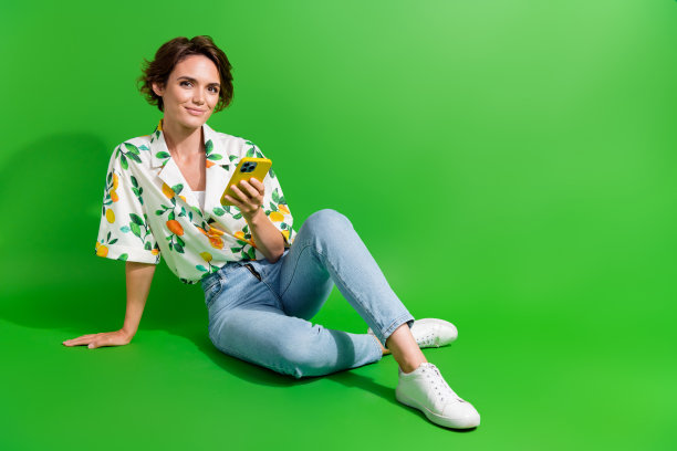 快乐的年轻女孩坐在地板上穿牛仔牛仔裤时髦的衬衫上网冲浪手机孤立的绿色背景的全长照片图片下载