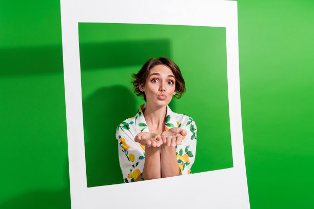 照片中，梦幻可爱的女孩穿着印花衬衫，拿着纸框，手伸着胳膊，亲吻着孤立的绿色背景图片下载