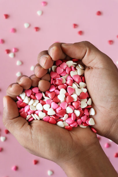 无法辨认的人捧起装满红色、粉色和白色心形糖果的手的特写图像，情人节的装饰，斑驳的粉色背景，抬高的视角，关注前景，情人节和浪漫的概念图片下载