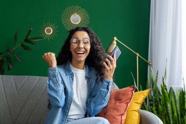 这张图片捕捉到一位快乐的女人在装饰时尚的房间里舒适地坐着，庆祝个人胜利或在智能手机上收到好消息图片下载