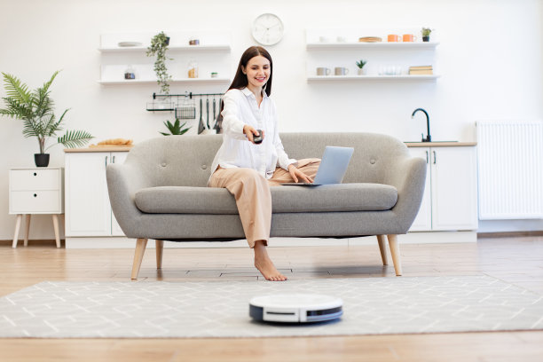 坐在沙发上的女士通过遥控器打开机器人吸尘器。图片下载