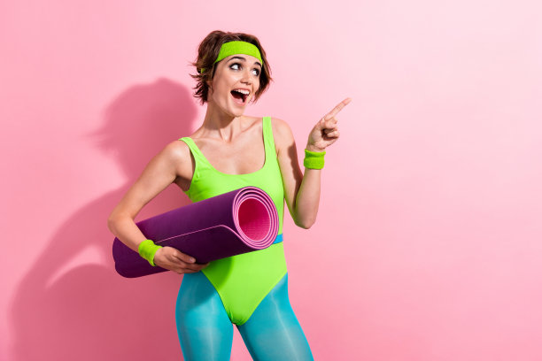 照片中漂亮的年轻女孩在体操垫上点了点空，穿着时髦的绿色运动连衣裤，孤立在粉红色的背景上图片下载