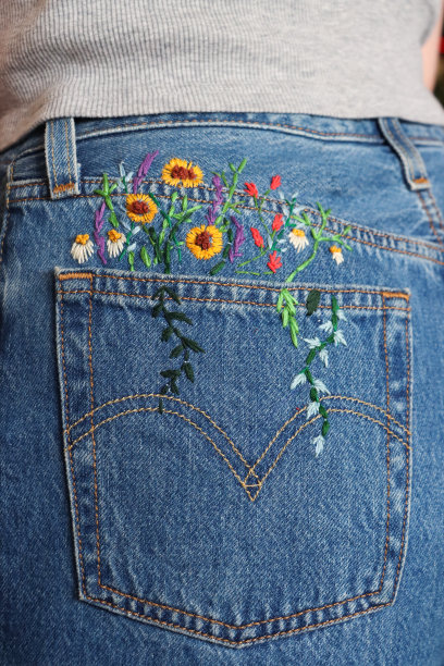 特写照片中，一个难以辨认的女人穿着自制定制的刺绣蓝色牛仔牛仔裤，刺绣的花朵和叶子，雏菊和向日葵从后口袋里长出来，后视图，聚焦于前景图片下载