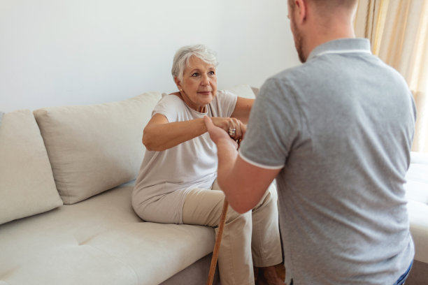 女居家照顾搀扶着老太太从沙发上站起身来。男性专业护理人员白天在家照顾老年妇女的照片。图片下载