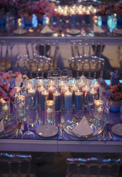 在昏暗的室内空间里，一张装饰着紫色花朵、插在玻璃罐里的紫色蜡烛、玻璃盘子、白色餐巾、红掌、兰花和山茶花的婚礼桌子。库存图片图片下载