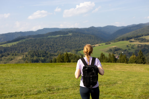 30多岁的女孩背着双肩包站在山顶上，绿草如草，在夏天日落的雾中看着美丽的山谷。风景与运动的年轻女子，后视图图片下载