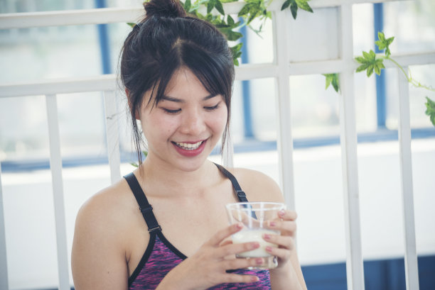 女人的手拿着白色的玻璃杯，牛奶从瓶子里倒出来。亚洲女性笑着笑着看镜头保健居家健身生活方式。美女饮用新鲜牛奶，低脂，无乳糖图片下载