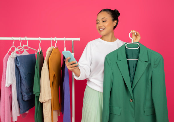 美丽的亚洲女人在衣架上拿着西装外套，在智能手机上浏览应用程序图片下载