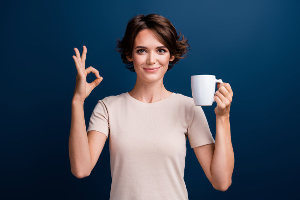 迷人的年轻女子的照片肖像拿着咖啡杯显示好符号穿着时髦的白色衣服孤立在深蓝色的背景图片下载