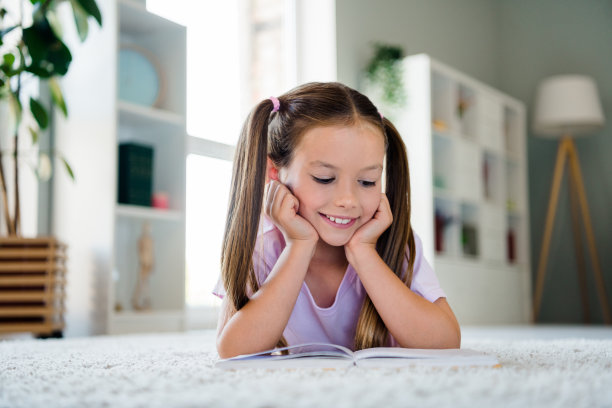 可爱的积极的小学生躺在地毯地板上的照片，双手触摸脸颊，享受阅读最喜欢的书图片下载