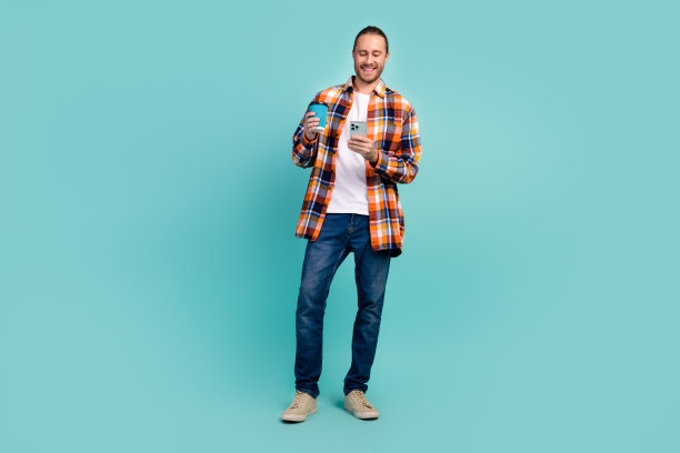 穿着法兰绒衬衫的酷男人的全身照片，拿着智能手机外卖杯，咖啡休息时间孤立在绿松石色背景上图片下载