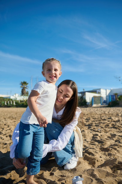 可爱的小女孩微笑着看着镜头，在温暖的阳光灿烂的日子里和妈妈在沙滩上玩耍图片下载