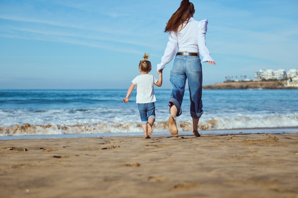 一位年轻的白人母亲和她的小女儿在沙滩上赤脚散步时手牵着手的背影图片下载