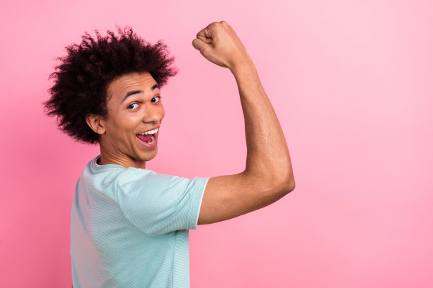 照片中的幸运酷男穿着蓝色t恤举起拳头，显示肌肉空白，孤立的粉红色背景图片下载