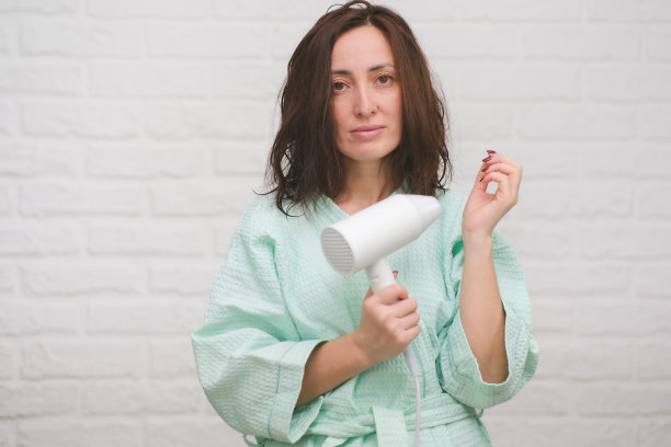 一个用吹风机微笑的漂亮女孩。微笑的女人在白色的桦树背景上用吹风机吹干头发。图片下载