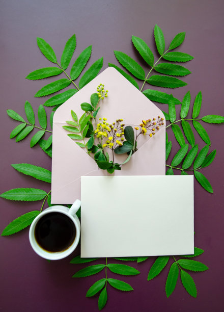 扁平的近距离粉色模型信封，里面装满了黄色的小花和小树叶，带有春天的绿叶，一杯咖啡在深紫色的背景上图片下载