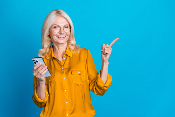 照片肖像迷人的退休妇女电话点空空间穿时髦的黄色丝绸衣服孤立在蓝色的背景图片下载