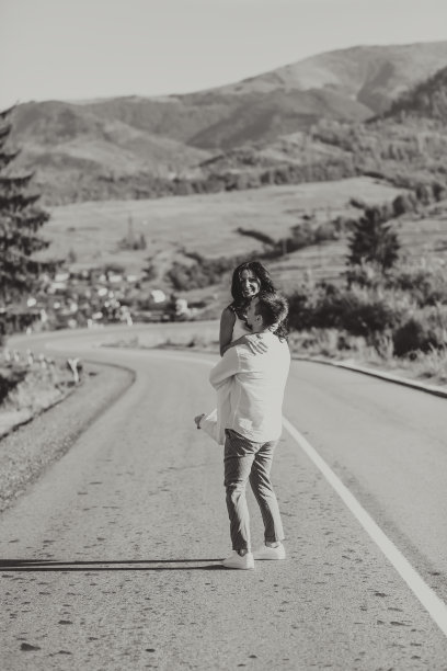 穿着白色衬衫的时髦新郎和身穿白色礼服的可爱黑发新娘在以森林和山脉为背景的柏油路上盘旋。黑白照片图片下载