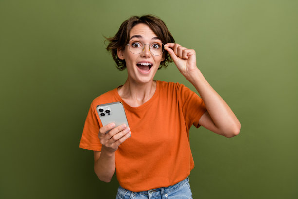 照片中，一个穿着橙色t恤的女孩喜出望外，拿着智能手机触摸眼镜，在卡其色的背景下进行销售图片下载
