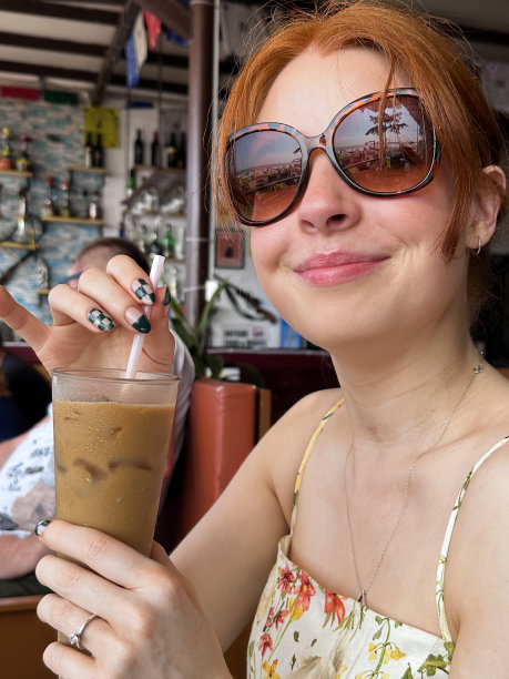 特写照片，迷人的红发女子坐在露天餐厅，拿着一杯加冰块的冰咖啡饮料，用吸管啜饮，在户外戴着太阳镜和背心裙，露天用餐的背景，重点在前景图片下载