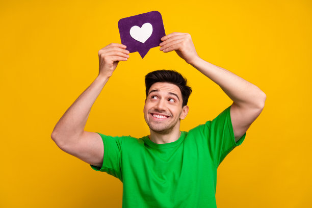 照片肖像英俊的年轻的黑发男子金色look心脏图标穿着时尚的绿色服装孤立在黄色的背景图片下载