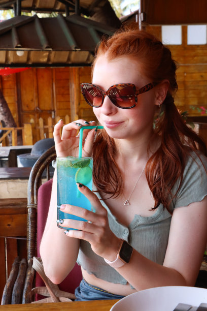 特写图片，迷人的红头发的女人啜饮蓝色泻湖伏特加和蓝色curaao鸡尾酒饮料与塑料吸管，冰块，柠檬片，薄荷叶，户外戴着太阳镜，户外用餐设置背景图片下载