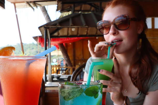特写照片中，迷人的红发女人用塑料吸管、冰块喝着彩虹模拟鸡尾酒，在户外戴着太阳镜，桌子上有蓝色泻湖和龙舌兰日出鸡尾酒，露天用餐背景图片下载