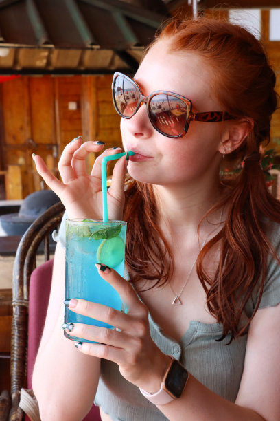 特写图片，迷人的红头发的女人啜饮蓝色泻湖伏特加和蓝色curaao鸡尾酒饮料与塑料吸管，冰块，柠檬片，薄荷叶，户外戴着太阳镜，户外用餐设置背景图片下载