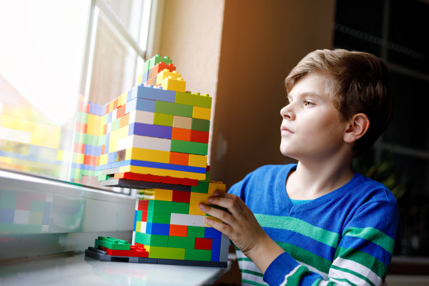 小男孩在玩许多五颜六色的塑料积木。可爱的小学生在窗户边玩建筑和创造建筑。创意休闲技术和机器人在日冕时间图片下载