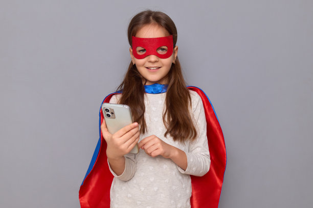 快乐、积极、开朗的小女孩穿着超级英雄的服装，戴着面具，孤立在灰色的背景下，拿着手机上网上网交流图片下载