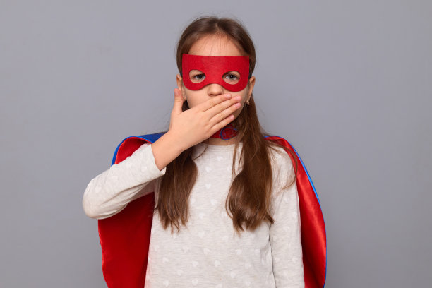 小女孩穿着超级英雄的服装，戴着面具，站在灰色的背景下，惊讶地看着，用手掌捂住嘴，看到了令人惊讶的事情图片下载