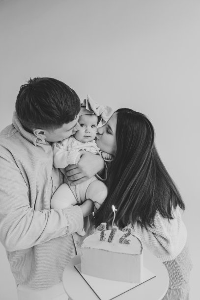 爸爸、妈妈牵着手逗小孩。创意生日蛋糕与蜡烛。家庭庆祝六个月大的婴儿生日聚会。黑白照片。爸爸妈妈亲吻隔离在墙上的可爱女孩图片下载