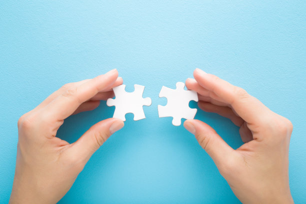 年轻的成年妇女的手指握住并连接不同的两个白色拼图片在浅蓝色的桌子背景。柔和的颜色。特写镜头。兼容性的概念。视角镜头。自顶向下视图。图片下载