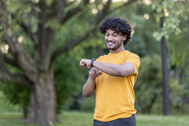 一个微笑的时尚印度男人在公园里做运动和跑步的照片。站起来，看着他手上的智能手表，检查脉搏，检测时间图片下载