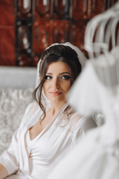 一位身穿晨衣的黑发新娘坐在灰色的沙发上，她的黑色卷发在一个穿着婚纱的人体模型后面，背景是粉刷过的墙壁。婚礼的照片。美丽的新娘图片下载