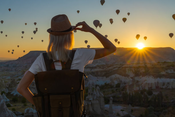 土耳其卡帕多西亚，一名年轻女子戴着帽子，背着手工背包，在山后冉冉升起的阳光下俯瞰气球，这张照片令人着迷。年轻的旅行者欣赏美丽的日出图片下载