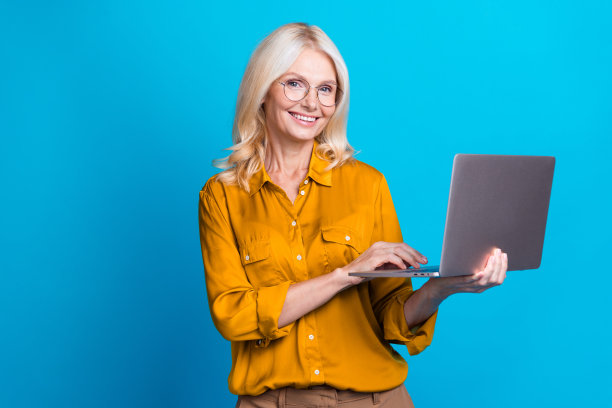 金发可爱的高级女士的照片拿着上网本编码游牧民族穿着时髦的黄色丝绸服装孤立在蓝色背景图片下载