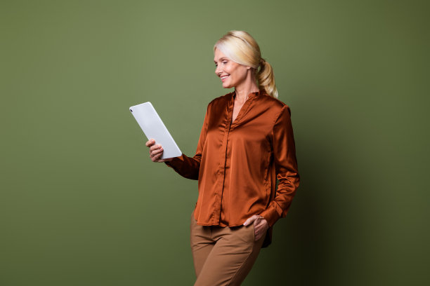 可爱的资深女士的照片肖像拿着阅读电子书穿着时尚的棕色丝绸服装孤立的卡其色背景图片下载