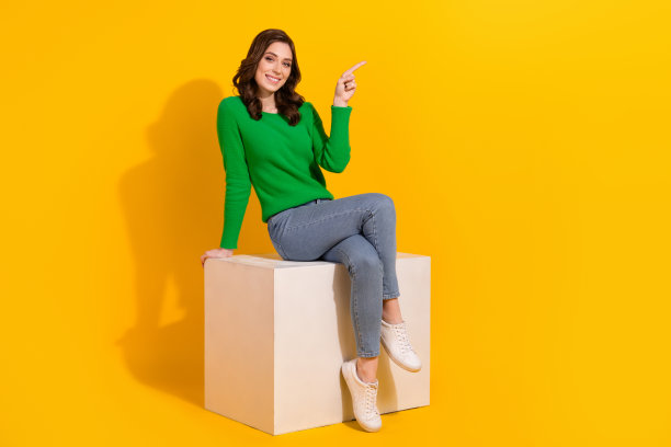 模特女性的全身长度照片，深色头发坐在讲台上，用手指点着黄色背景，使她的机构孤立起来图片下载