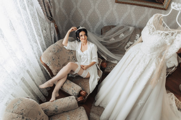 身穿晨衣的黑发新娘躺在灰色沙发上为镜头摆姿势。广角照片。优雅的发型。漂亮的化妆品。拖地长裙。新娘的早晨。图片下载
