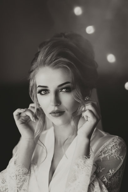 黑白照片。一个金发碧眼的新娘穿着一条长长的蕾丝衬裙，站在她的房间里，在镜子前摆姿势，戴上耳环。漂亮的发型和妆容，敞开胸围。婚礼的肖像。镜子里的倒影图片下载