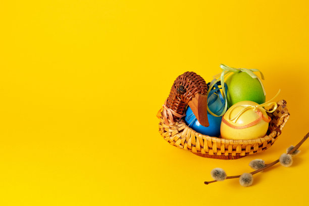 手工鸭形稻草篮子，五颜六色的彩蛋，缎面蝴蝶结和缎带和柳枝。黄色背景的横幅。副本的空间。空文本位置。复活节卡片。内部装饰图片下载