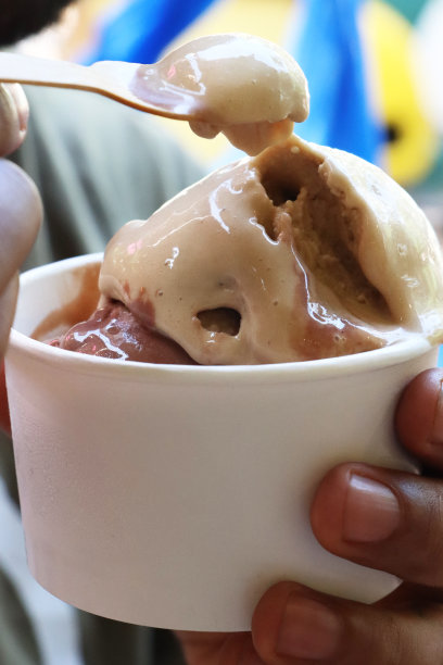 特写图片:一个不知名的人拿着木勺，装着巧克力和香草奶油的白色一次性纸板桶，焦点在前景图片下载