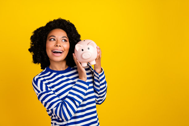 照片中开朗好奇的女子穿着条纹运动衫，摇晃着钱猪盒子，在黄色背景上显得空荡荡的孤立图片下载