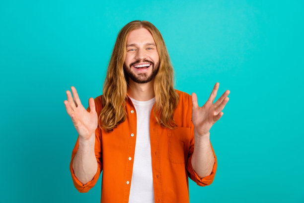 年轻的嬉皮士男子的照片金发长发英俊的模特在橙色衬衫张开双臂时高兴地看到你孤立的青色背景图片下载