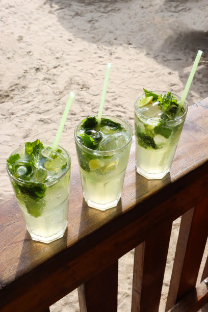 在露天海滩酒吧餐厅的木栏杆上，一排三杯鸡尾酒的图片，莫吉托鸡尾酒的玻璃杯，冰块，薄荷叶枝和酸橙片，吸管，沙子背景，高架视图，重点在前景图片下载