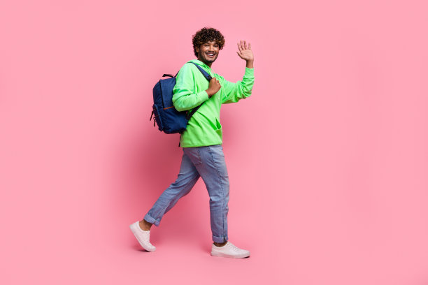 微笑的年轻友好学生的全身照片，背着背包，在粉红色的背景下向观众挥手图片下载