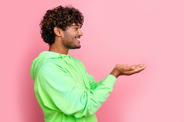 侧面侧面照片的开朗的年轻人在绿色运动衫拿着两只手的广告最喜欢的品牌孤立在粉红色的背景图片下载