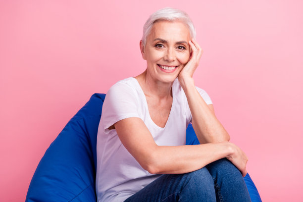 照片中，长着牙齿、笑容满面的好心情的女人留着短发，穿着白色的t恤，手掌放在颧骨上，坐在粉红色的背景上图片下载